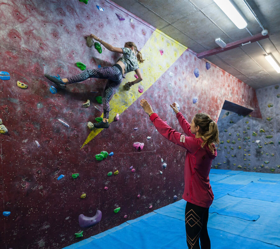 A coach has arms in the air to spot a boulderer who is climbing on a wall