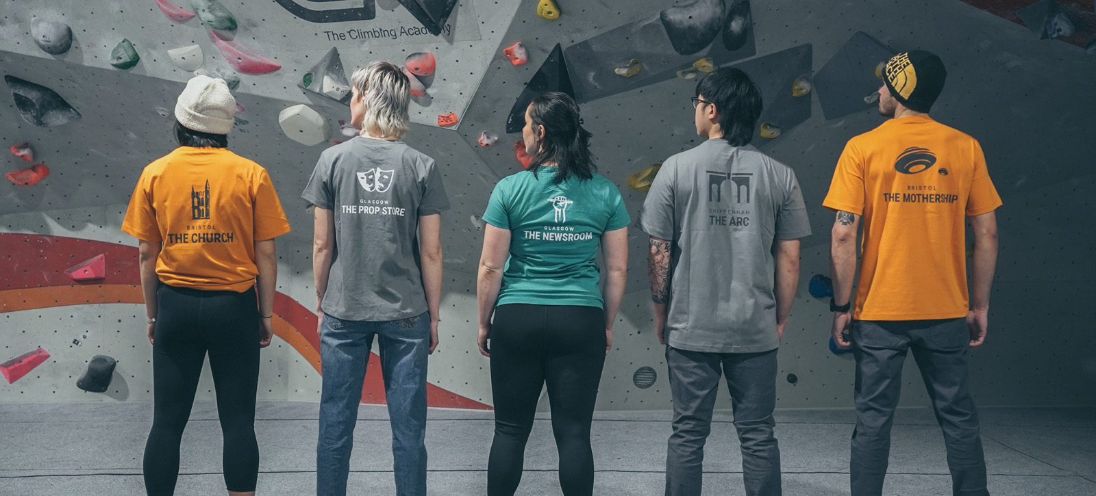 5 climbers (mixed mean and women) standing with their backs to us, in front of a climbing wall, wearing t-shirts that are branded with each of the TCA climbing centres.