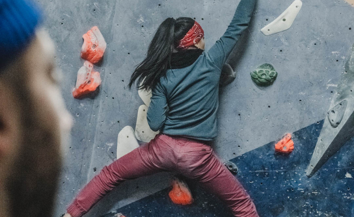 A woman in a head band is shown from behind whilst bouldering. We see her arms and legs outstretched