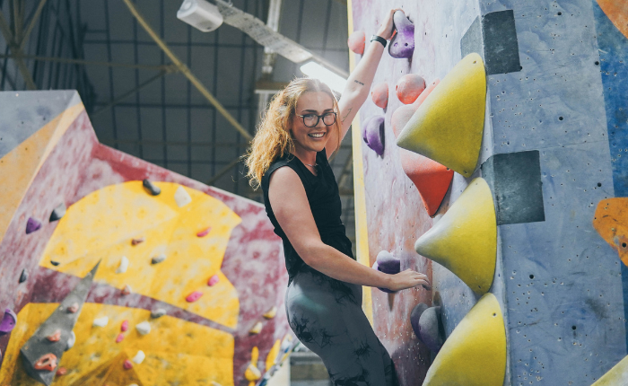 A woman smiles at the camera while hanging onto the wall bouldering