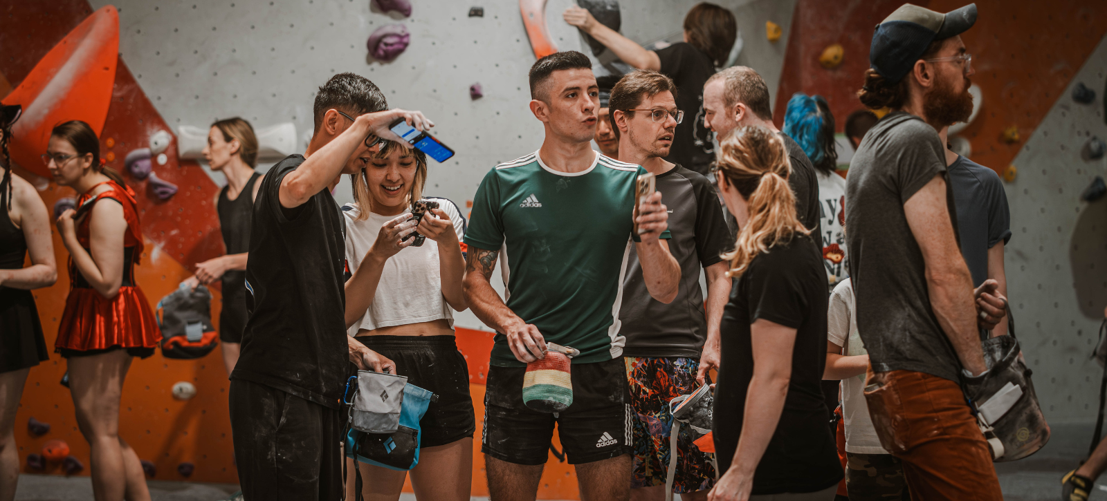 A mixed group of climbers stand on the bouldering mats chatting, laughing and looking at a phone