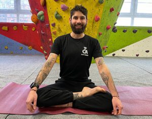 Instructor Jack sits in a cross-legged yoga pose on a mat, with a climbing wall in the background. 