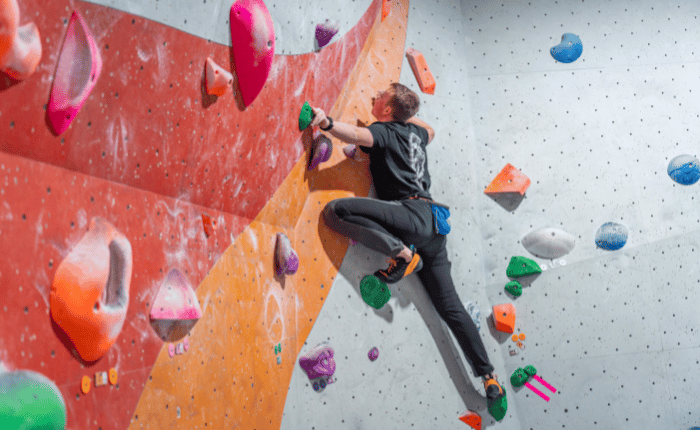 A man is bouldering with his back to us. His left foot is stepping up high and he is reaching out left to balance