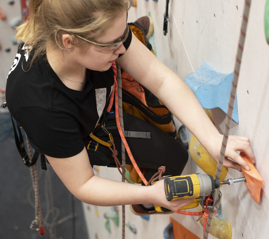 A female routesetter is screwing a climbing hold onto a wall whilst supported from a rope.