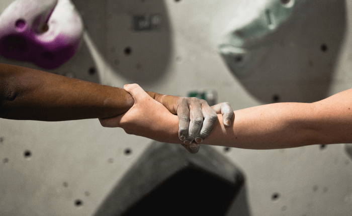 A person of colour holds hands with a white person while climbing the buddy challenge. We on;t see their hands and a climbing wall in the background.