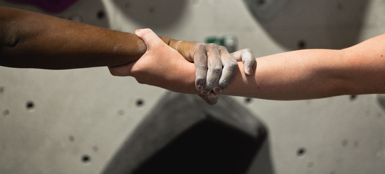 A person of colour holds hands with a white person while climbing the buddy challenge. We can see their hands and a climbing wall in the background.
