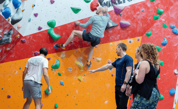 A mixed group gather around a boulderer, while pointing at and talking about other climbs