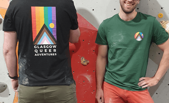 Two climbers sporting Glasgow Queer Adventures t-shirts. We see their torsos, one facing us smiling and one facing away from the camera.