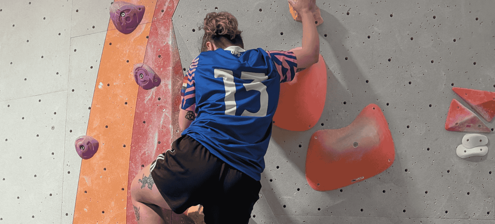 A boulderer is seen from behind. We can see the edge of a pride flag on their top.