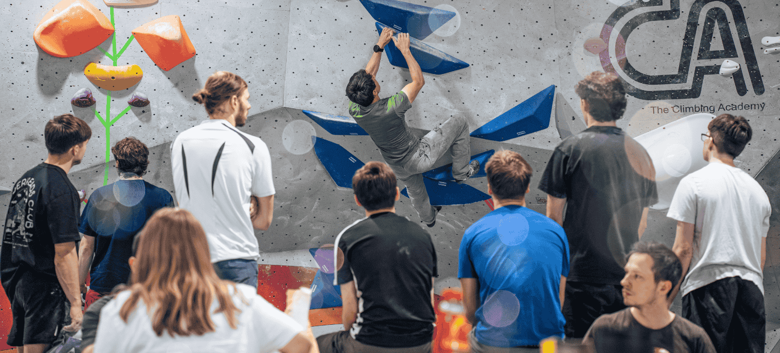 Climbers gather round a climber on a blue problem at a comp to await their turn