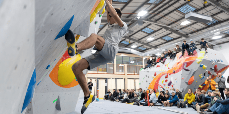 We see a climbing bouldering in the foreground with a crowd watching from the comfort of the the mats or sitting on top of the top-out boulder 