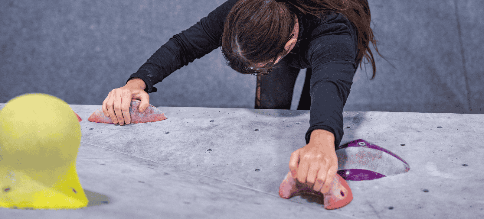 We see a climber from above gripping holds and looking down as they move up a boulder problem