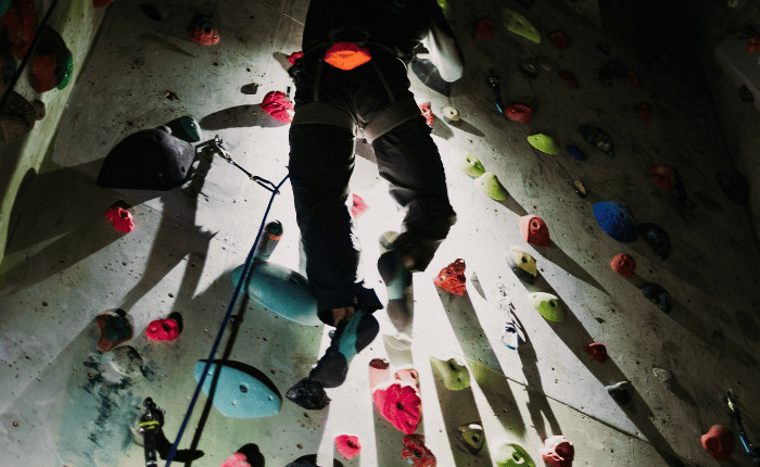 A climber in a dark room looks down while roped climbing leaving cool shadows protruding below him.