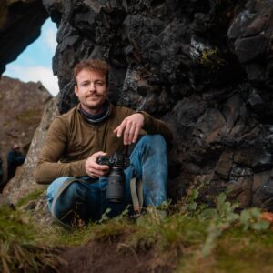 Ewan crouches with his camera in his hand by some rocks at Dumby