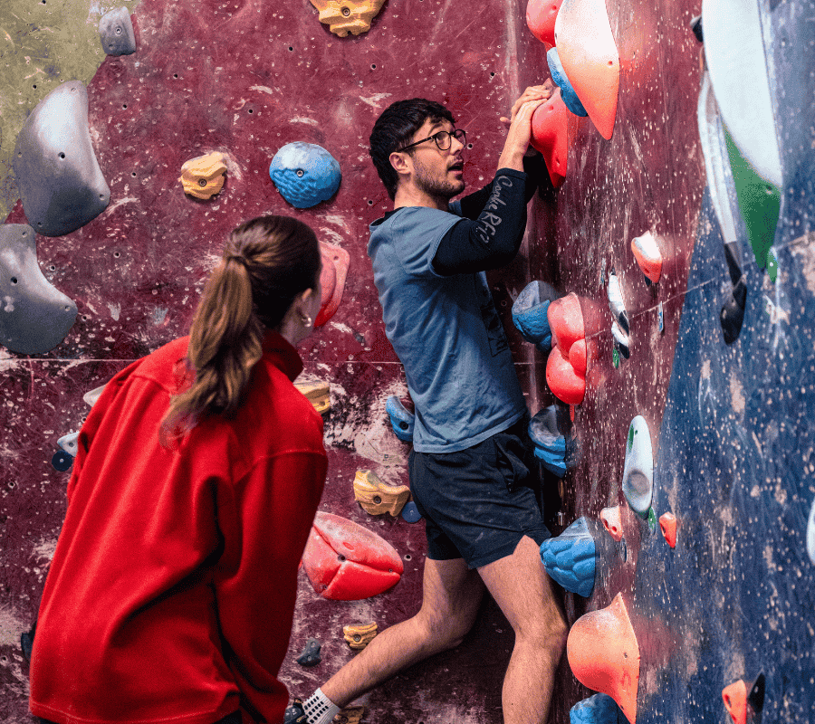 A woman encourages her climbing partner who is on the wall, looking up at his next move