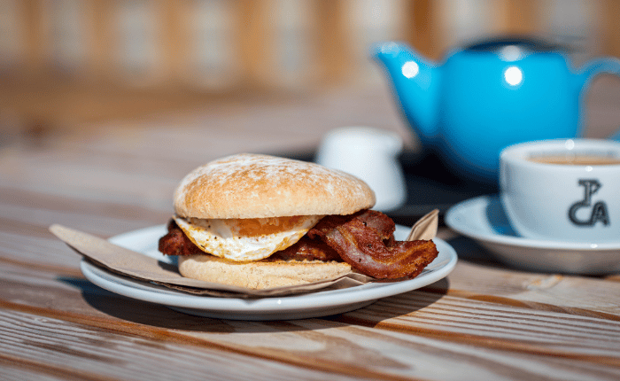 A breakfast bap with visible egg and bacon, a cup of tea and pot.
