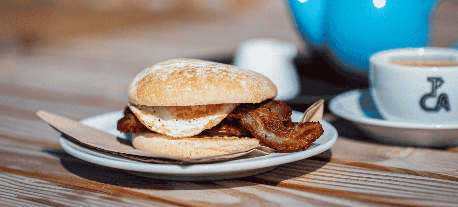 A breakfast bap with visible egg and bacon, a cup of tea and pot.