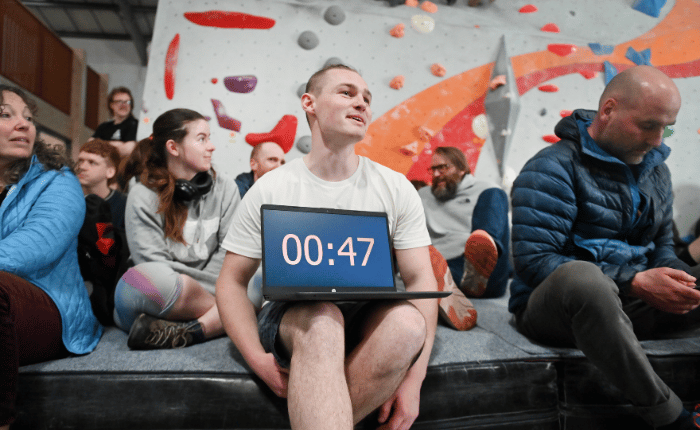 A smiling TCA staff member holds a timer display during the finals of a bouldering comp - they are seated on the mats surrounded by spectators.