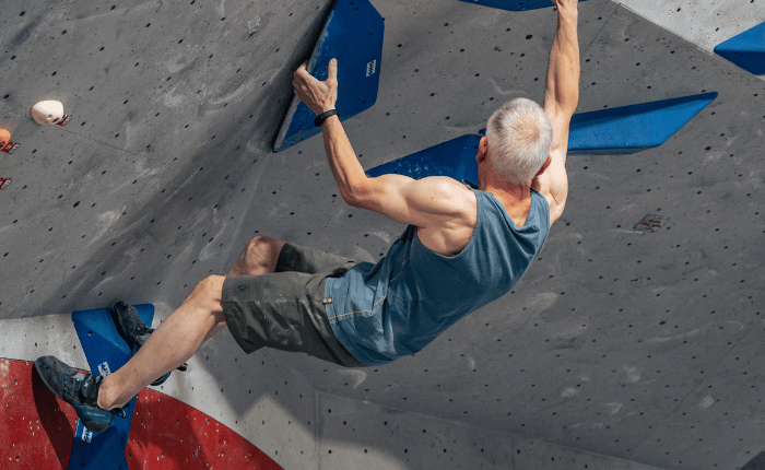 A man with grey hair and well defined muscles is seen bouldering from behind
