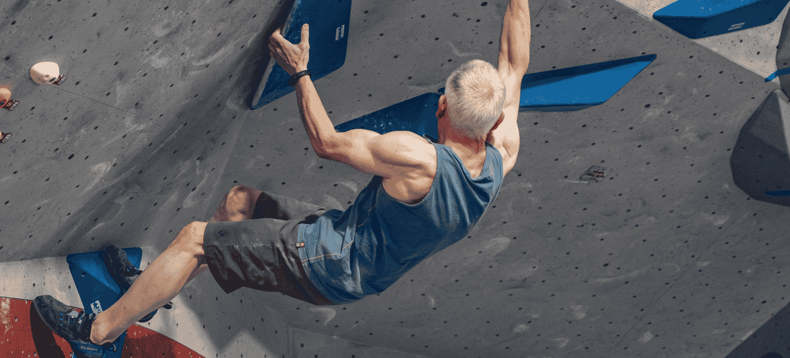 A man with grey hair and well defined muscles is seen bouldering from behind