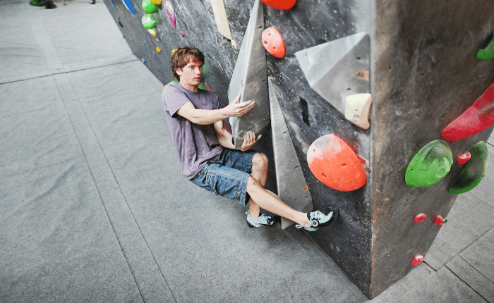 A boulderer holds himself off the ground by gripping an angular volume (whilst low over mats) with his focus on the next move.