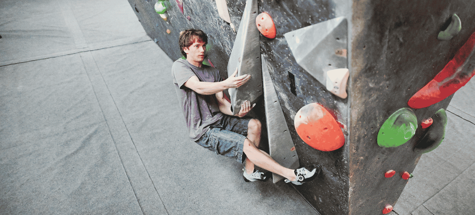 A boulderer holds himself off the ground by gripping an angular volume (whilst low over mats) with his focus on the next move.